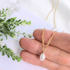 Gold chain necklace with a pearl pendant held between fingers on a white background with greenery.